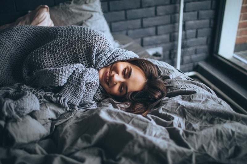 Women snuggled in a grey knit blanket on her bed.