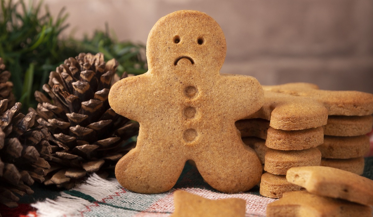 Gingerbread cookie with a sad face, next to a stack of other cookies set on a table with a pinecone and greenery for decor.