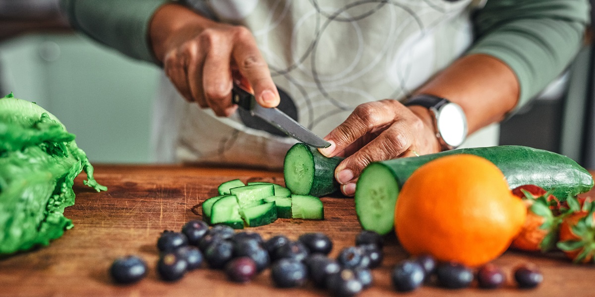 Man slicing up cucumber and other veggies on dark wood cutting board.