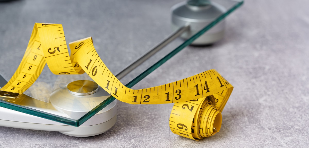 Yellow measuring tape curled over a glass floor scale on a grey floor.