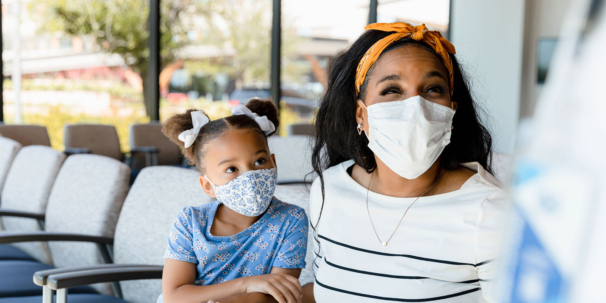 A mother and daughter wearing medical masks sit in the waiting room of an urgent care and are greeted by a nurse